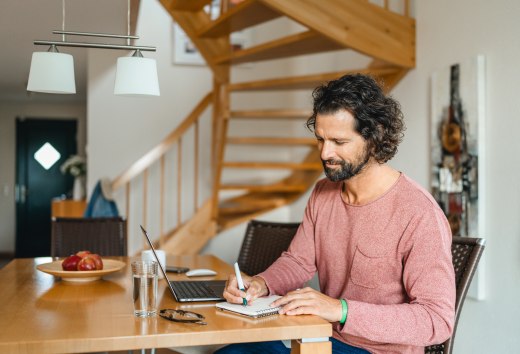Workation na wyspie Poel: pracuj wydajnie w przytulnym domu wakacyjnym i ciesz się wolnym czasem., © TMV/Gross Mężczyzna siedzi przy drewnianym stole w domku letniskowym, pracując na laptopie i robiąc notatki - idealne warunki do pracy na wyspie Poel.