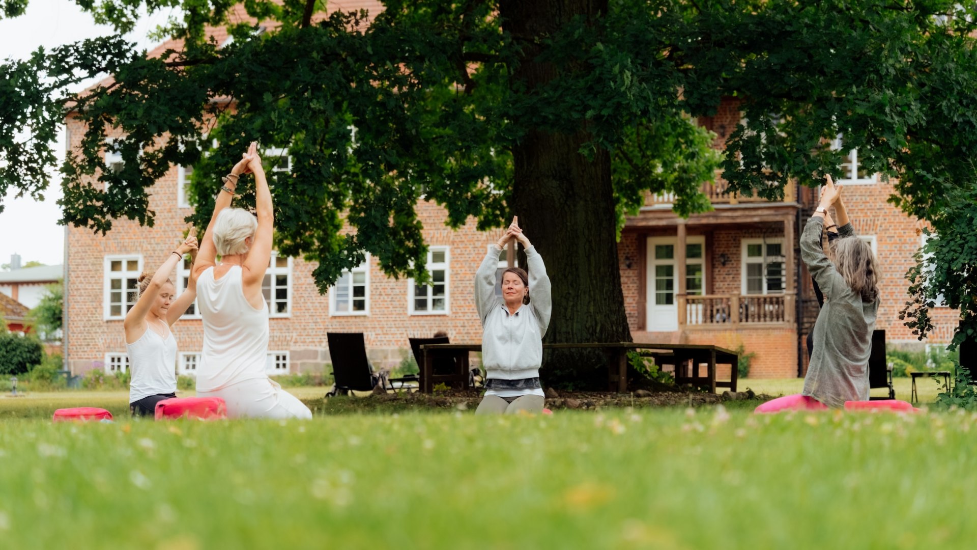 Three women are doing yoga in a meadow under a large tree, with the Ludorf manor house in the background. // Beneath an old oak tree, gentle movements flow through Ludorf Manor Park—yoga on the lawn, with the brick building in view, clearing the mind // © MV-T/Petermann Three women are doing yoga in a meadow under a large tree, with the Ludorf manor house in the background.