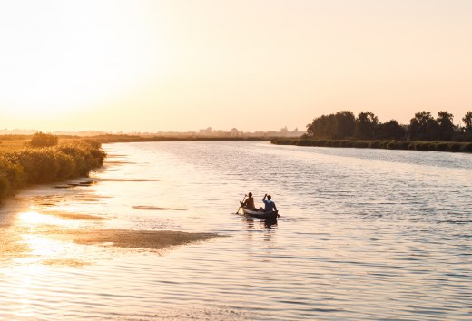Two people paddle in a canoe across a calm river at sunset, with golden light on the water.