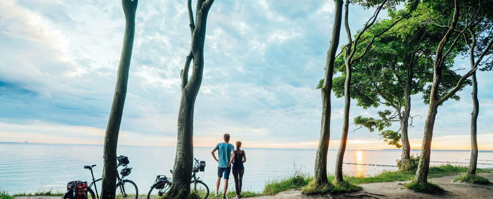 Cycle along the Baltic Sea coast in Mecklenburg-Vorpommern through the ghostly forest., © TMV/Gänsicke A couple stands in the ghostly forest, their bicycles leaning against the trees, watching the sun set over the Baltic Sea.