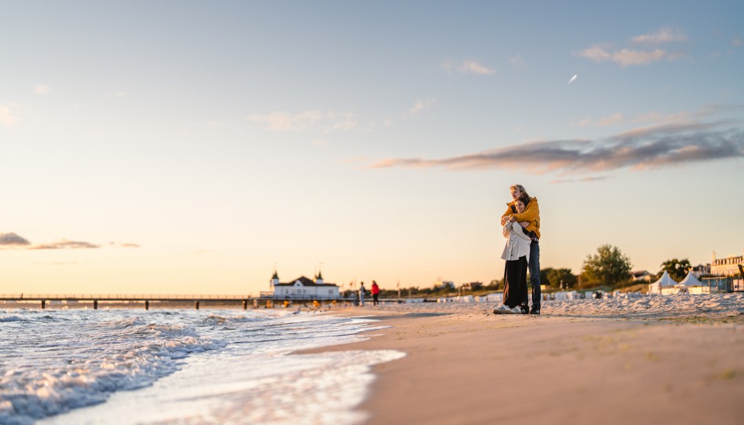Een stel staat elkaar omhelzend op het strand van Ahlbeck, met de pier en de avondhemel in warm licht. // Zonsondergang op het strand van Ahlbeck: samen langs het water wandelen, de uitgestrektheid voelen, even alles loslaten. // © MV-T/Gross Een stel staat elkaar omhelzend op het strand van Ahlbeck, met de pier en de avondhemel in warm licht.