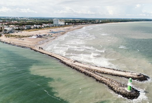 Luchtfoto van het strand van Warnemünde met pier, vuurtoren en de Baltische Zee in een zachte deining. // De Baltische Zee kabbelt in zachte golven tegen het brede zandstrand van Warnemünde. De stenen pier leidt naar zee, bekroond door de groen-witte vuurtoren. Strandstoelen staan in een rij langs de kust, terwijl het silhouet van de badplaats met zijn historische gebouwen en moderne hotels erachter te zien is. // © MV-T/Scholz-Witzel Luchtfoto van het strand van Warnemünde met pier, vuurtoren en de Baltische Zee in een zachte deining.