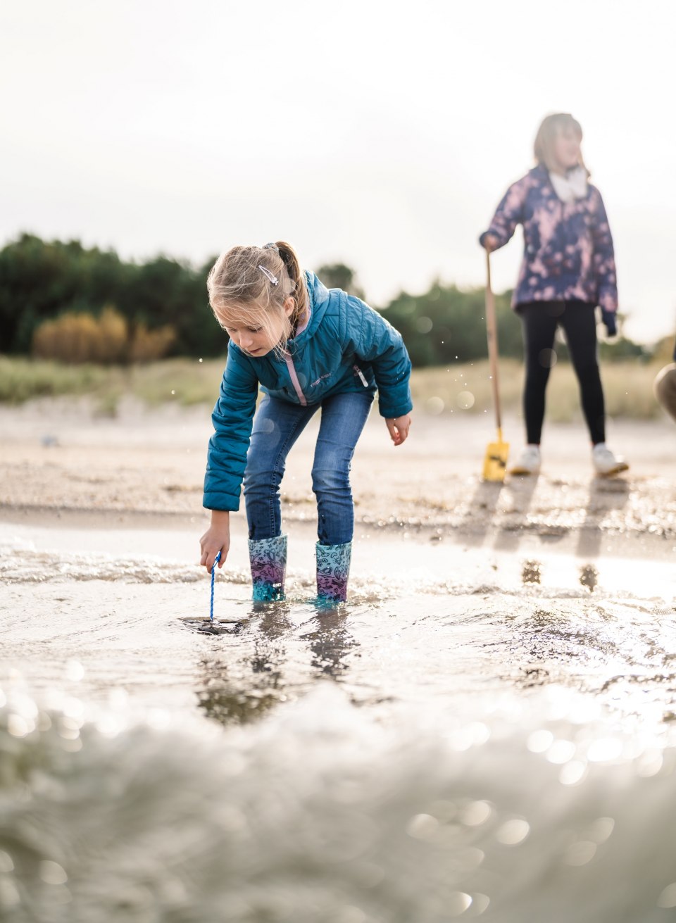 Een meisje in regenlaarzen staat voorovergebogen in het water op het strand, op zoek naar iets in het zand, terwijl een ander kind op de achtergrond staat.