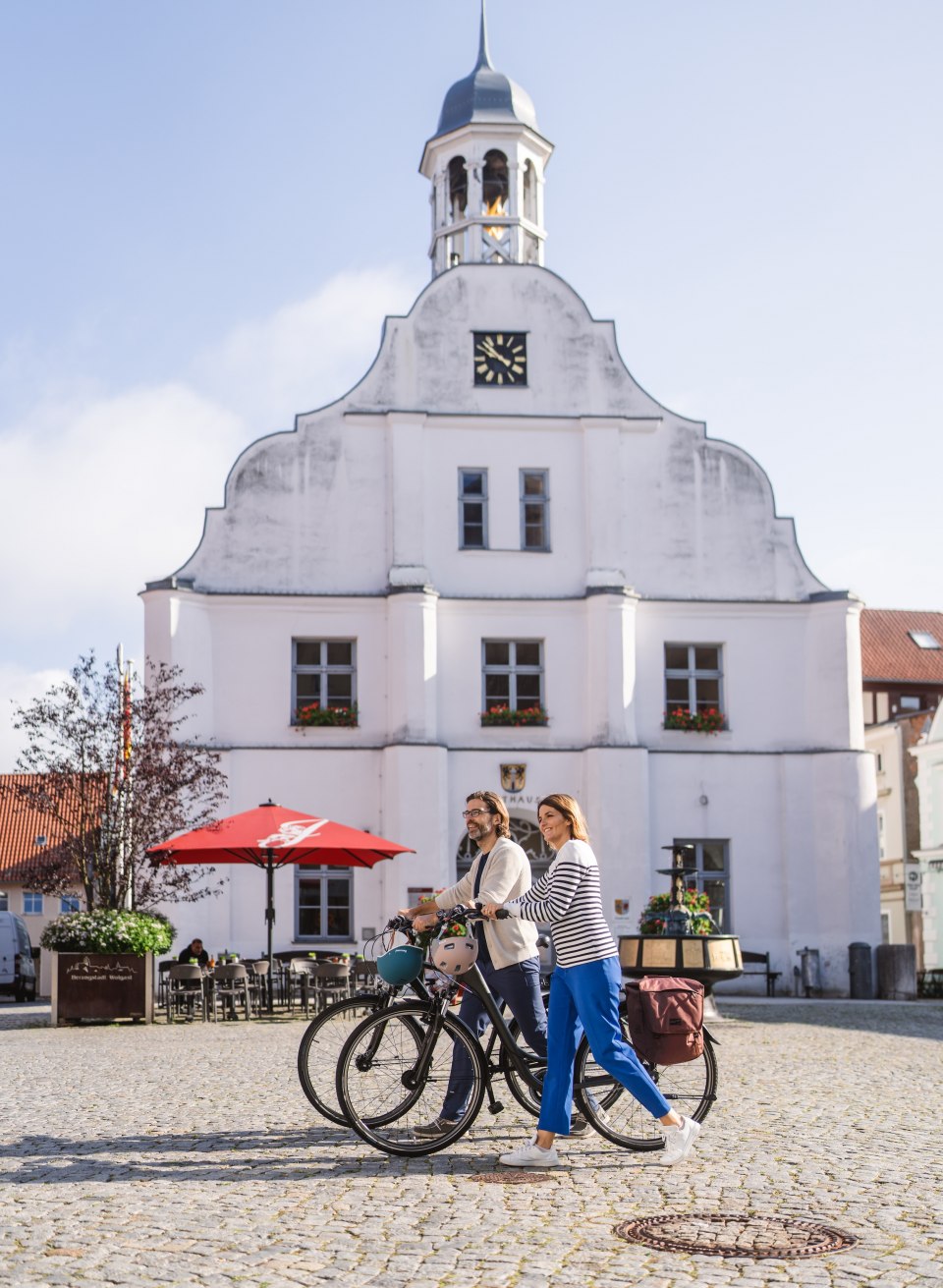 Two cyclists push their bikes across the market square in Wolgast // The market square in Wolgast is dominated by the gleaming white town hall. // © MV-T/Gross Two cyclists push their bikes across the market square in Wolgast