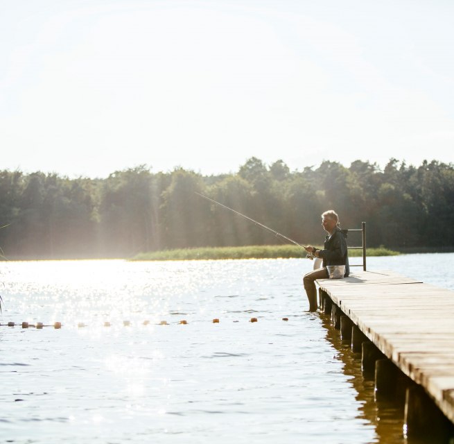 A man sits on a wooden jetty on a calm lake and fishes while the sun makes the water glisten.