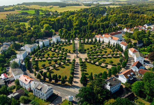 Luchtfoto van het Circus in Putbus op het eiland R&uuml;gen, een cirkelvormig plein met symmetrisch geplaatste bomen en classicistische gebouwen, omgeven door groene natuur en een landelijk landschap.