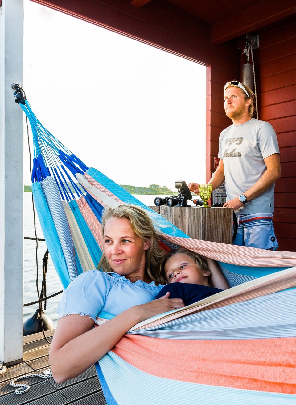 A woman and a child relax in a colorful hammock on a houseboat while a person at the helm looks out over the lake.