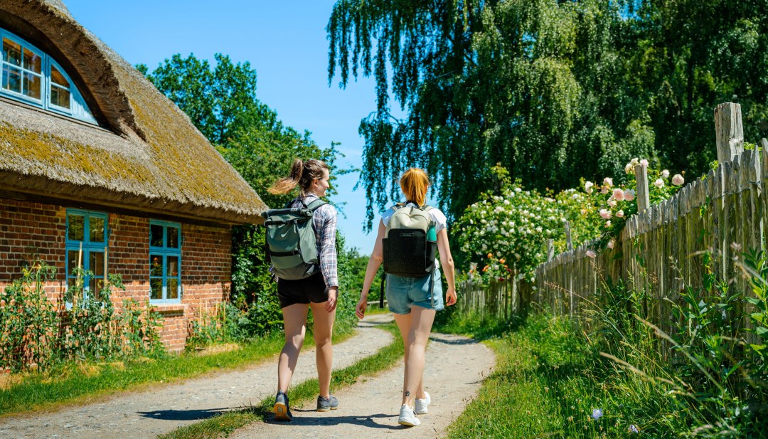 Twee jonge vrouwen lopen in de zon langs een huis met rieten dak en bloeiende tuinen in een idyllisch dorpje.