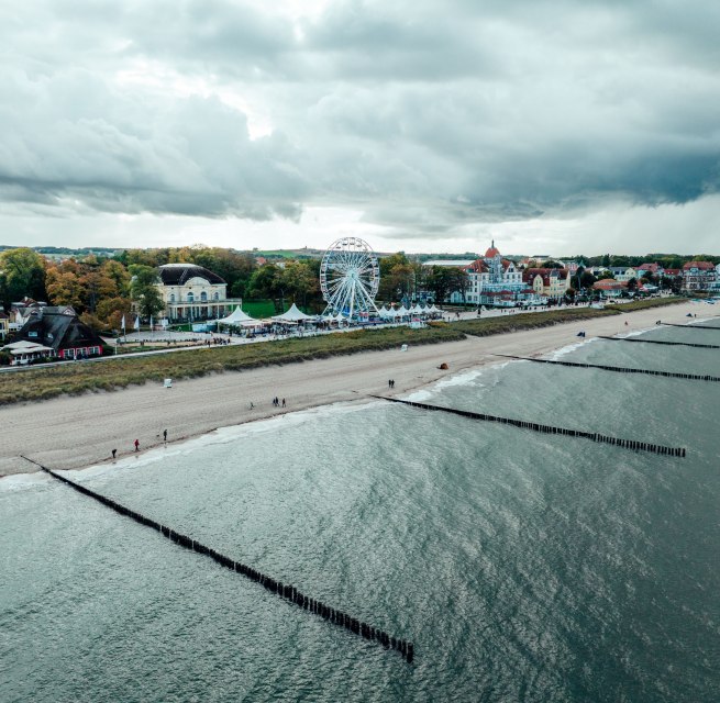 Luchtfoto van de strandpromenade van Kühlungsborn aan de Baltische Zee met zandstrand, strandhoofden, reuzenrad en historische gebouwen op de achtergrond.