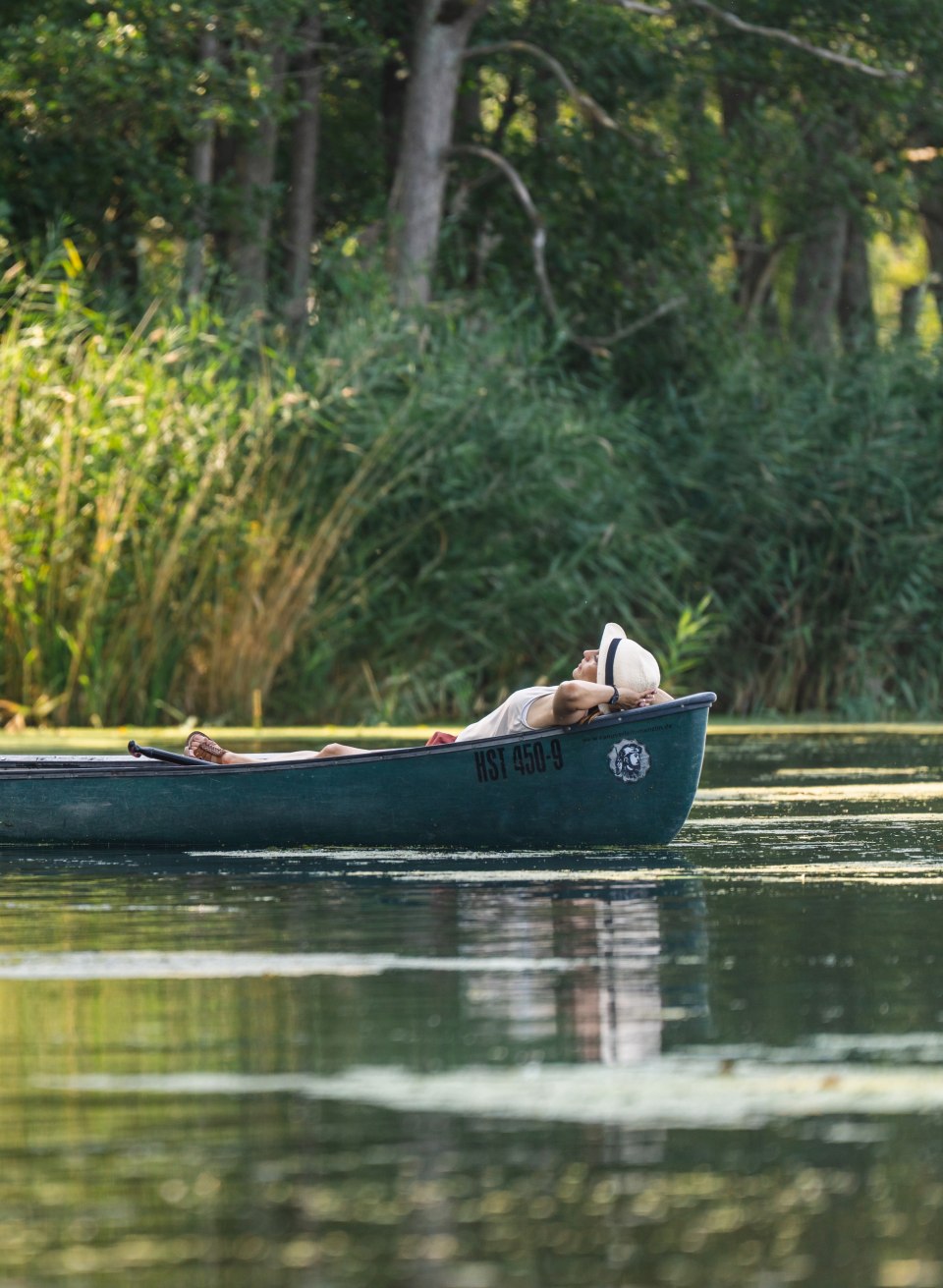 Two people are floating in a canoe on the Peene, one sitting ready to paddle, the other relaxing in the shade of the riverbank vegetation.