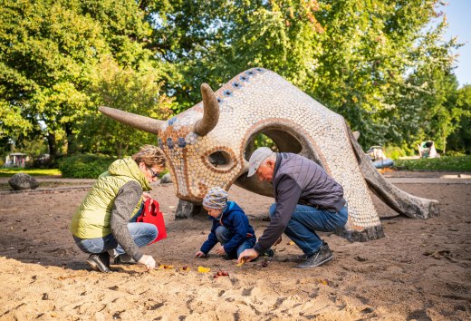 Eine Frau und ein Mann hocken mit einem Kind im Sand vor einer gekachelten Bullenskulptur und suchen Kastanien. // Kastanien im Sand, Laub in der Hand: Am Spielplatz am Tollensesee in Neubrandenburg sammeln Familien gemeinsam den Herbst auf. // © MV-T/Tiemann Eine Frau und ein Mann hocken mit einem Kind im Sand vor einer gekachelten Bullenskulptur und suchen Kastanien. // Kastanien im Sand, Laub in der Hand: Am Spielplatz am Tollensesee in Neubrandenburg sammeln Familien gemeinsam den Herbst auf. // © MV-T/Tiemann
