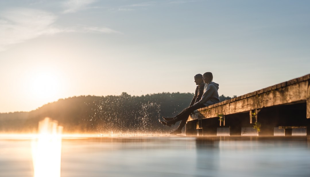 Two people sit on a jetty on Lake Mirov at sunrise and dip their feet into the smooth water while a fine mist hovers over the surface.