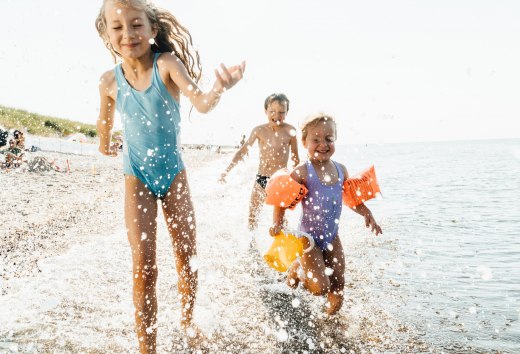 Three children run through the shallow water on the Baltic Sea beach, laughing and splashing happily in the waves. // Carefree bathing fun - laughing children romp on the beach and enjoy a summer's day at the Baltic Sea. // © MV-T/Roth Three children run through the shallow water on the Baltic Sea beach, laughing and splashing happily in the waves.