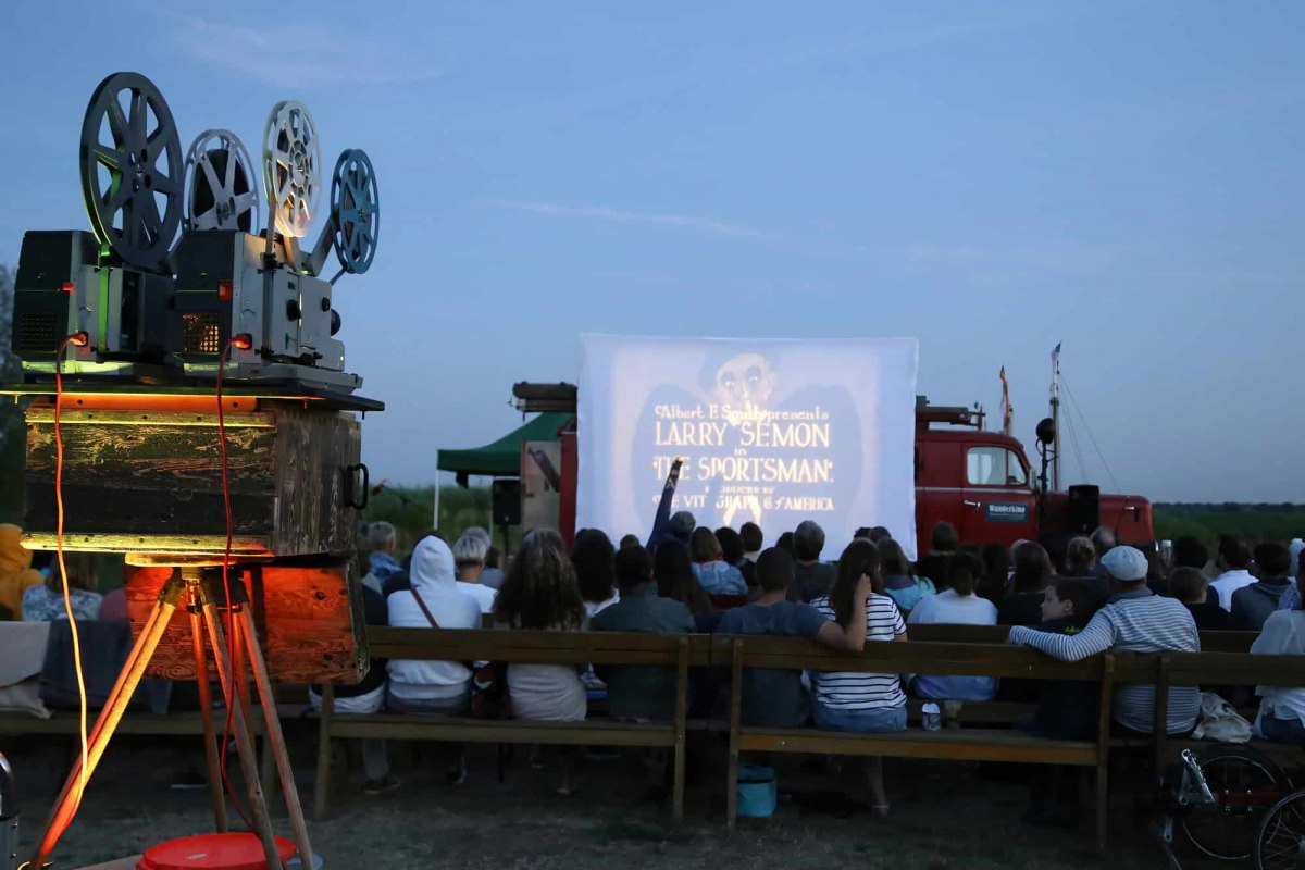 An open-air movie night hosted by the Althagen Traveling Cinema in Ahrenshoop, featuring a historic 16mm or 35mm film projector in the foreground and an audience seated in front of the screen. // © Tobias Rank An open-air movie night hosted by the Althagen Traveling Cinema in Ahrenshoop, featuring a historic 16mm or 35mm film projector in the foreground and an audience seated in front of the screen. // © Tobias Rank