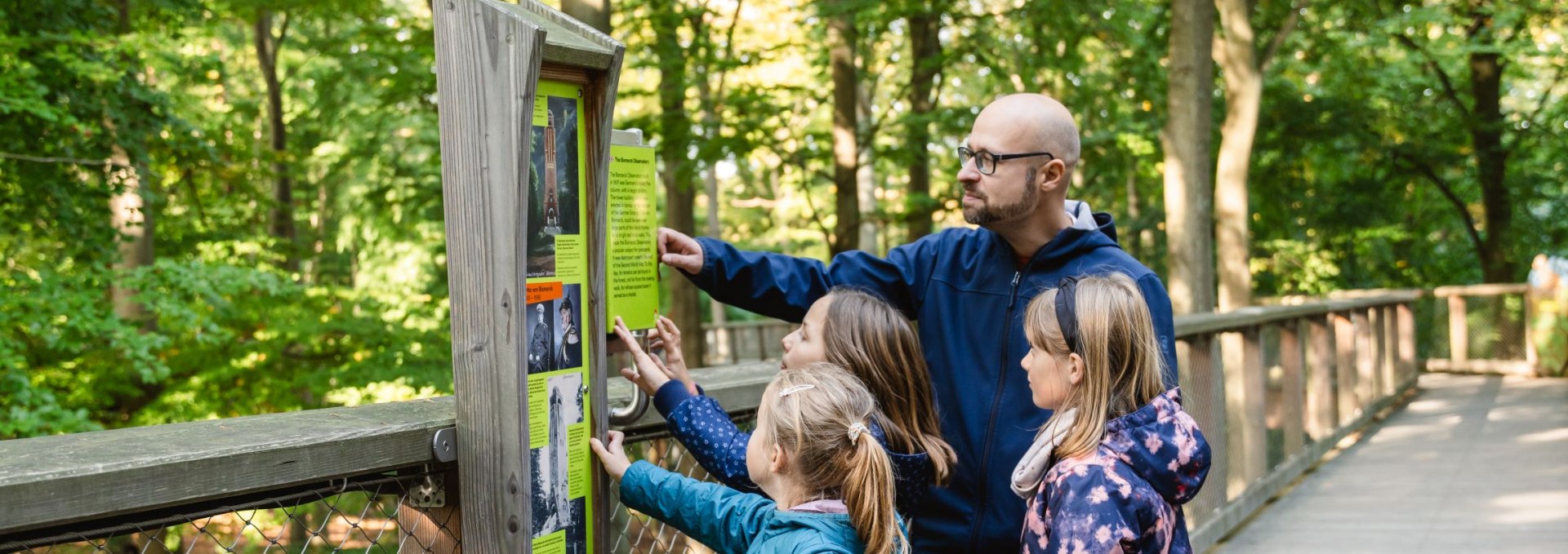 Een groep kinderen en een volwassene bekijken een informatiebord op een houten boomtoppenpad omringd door groene bomen.