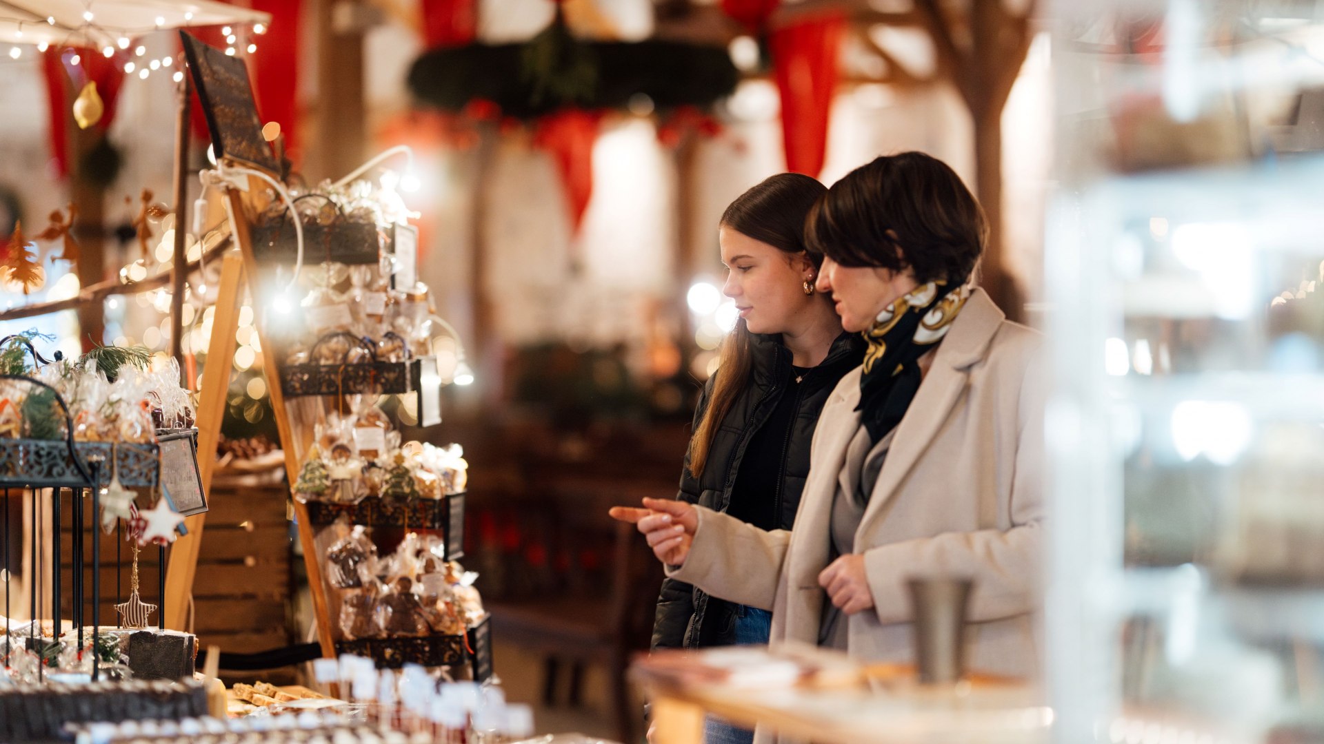 Twee vrouwen wandelen over een sfeervol verlichte kerstmarkt en bekijken kunstig ingepakt snoep en versieringen.