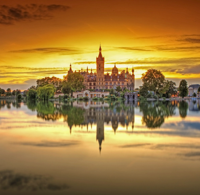 Kasteel Schwerin wordt omlijst door de laatste zonnestralen. De zonsondergang kleurt de lucht oranje. Kasteel Schwerin wordt weerspiegeld in het kalme meer.