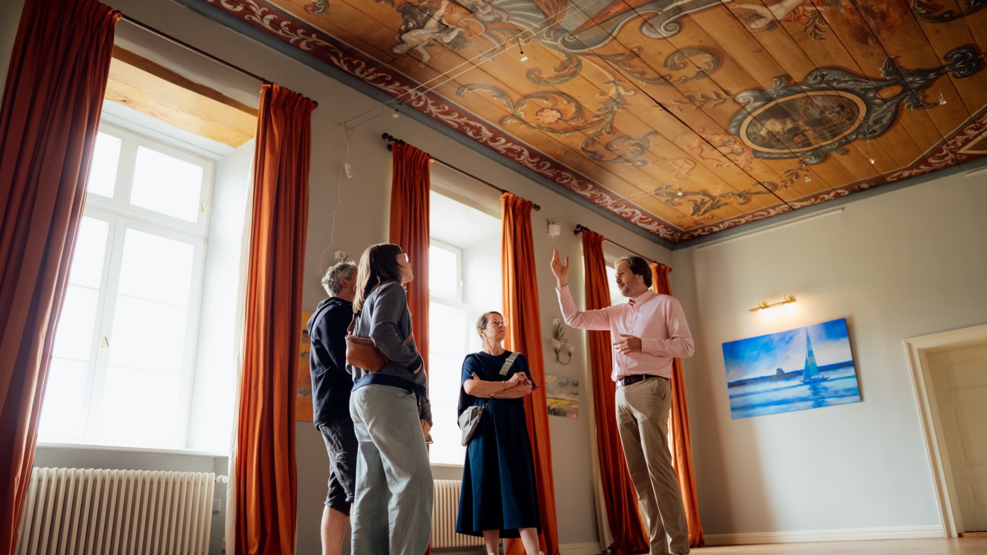 Jannis Achtenhagen explains the painted ceiling and the tall windows with curtains to guests in the Baroque Hall of the Ludorf Manor House. // Annis Achtenhagen raises her hand, directs everyone’s attention to the painted ceiling, and shares the next details—a guided tour of the Baroque Hall at the Ludorf Manor House // © MV-T/Petermann Jannis Achtenhagen explains the painted ceiling and the tall windows with curtains to guests in the Baroque Hall of the Ludorf Manor House.