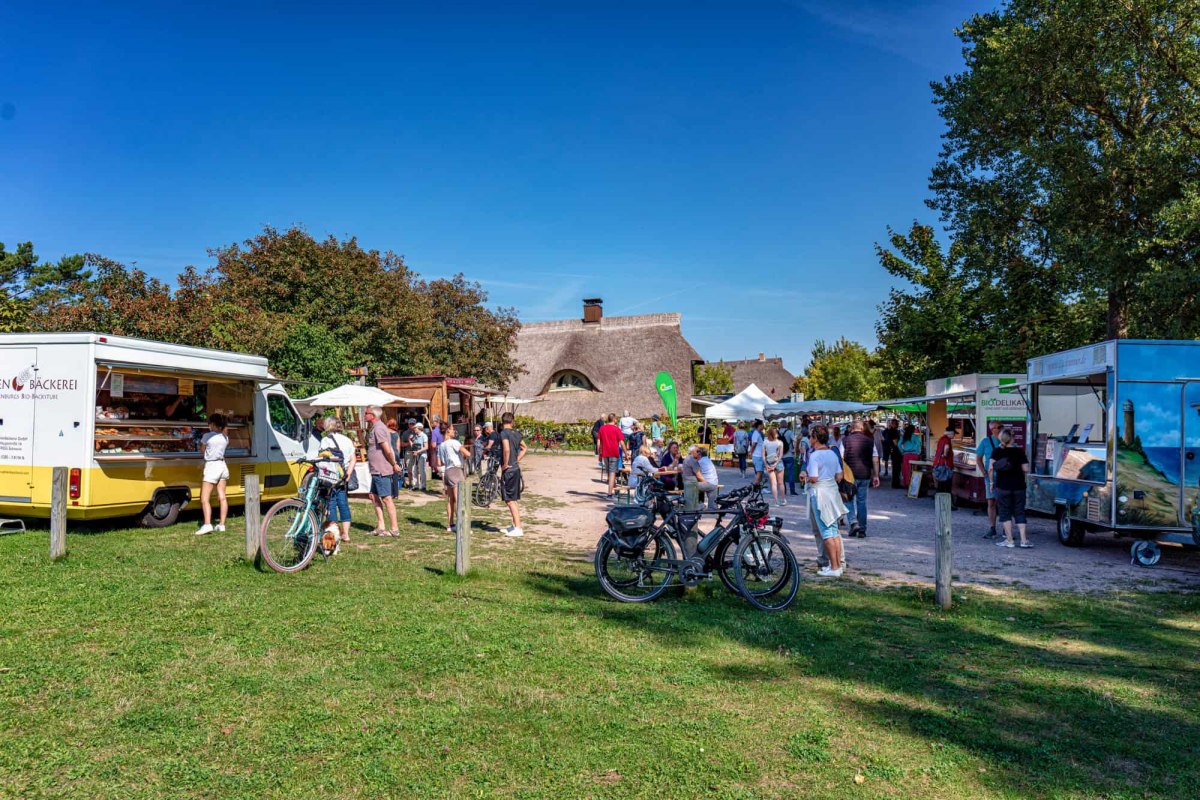 De zomermarkt van Ahrenshoop nodigt je uit om te snuffelen en te genieten in een ontspannen sfeer. Tussen traditionele rietgedekte huisjes presenteren regionale handelaren een breed scala aan producten op hun trailers - van verse bakkerswaren tot biologische lekkernijen. Op de foto zie je de levendige drukte buiten, waar bezoekers op de fiets aankomen en aan kleine tafeltjes blijven hangen om de typische flair van de Oostzeeregio te ervaren. // &copy; Fa. Voigt & Kranz UG, Prerow