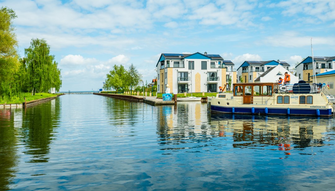 The adventure begins: Cautiously and full of anticipation, the first houseboat leaves the protected marina in the harbor village of Müritz (Rechlin)., © TMV/Tiemann A houseboat leaves a canal towards the lake and in the background you can see houses with vacation apartments.