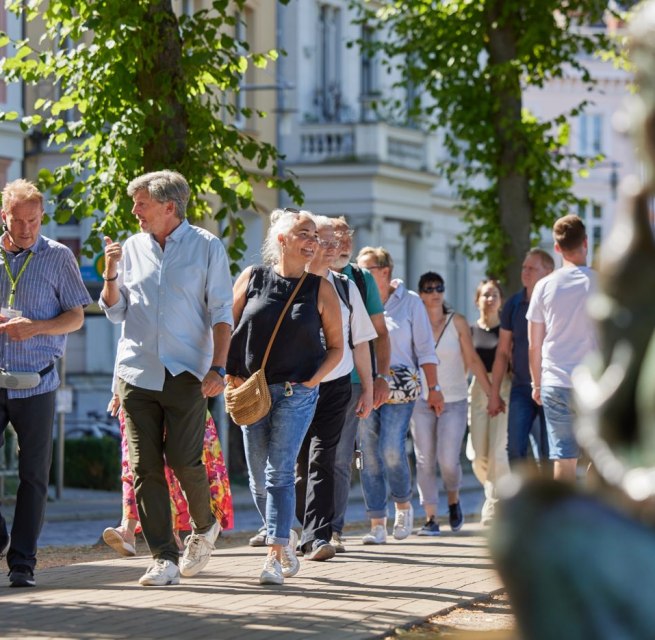 A group of people are walking along a cobbled path with a city guide. On the right, blurred in the picture, a statue can be seen by the Pfaffenteich pond; the villas appear in the background. The focus is on the guide and a couple having a lively conversation., © Oliver Borchert