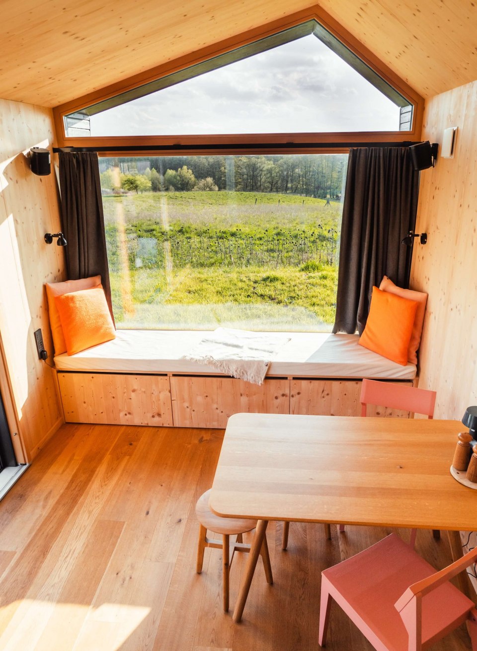 Stijlvol minimalisme ontmoet natuur - tiny house met panoramische ramen en gezellig interieur voor een ontspannen vakantie., © mari.linni || Nicola Link Berlin Licht Tiny House met groot raam aan de voorkant, houten interieur en bank met uitzicht op een weids, groen landschap.