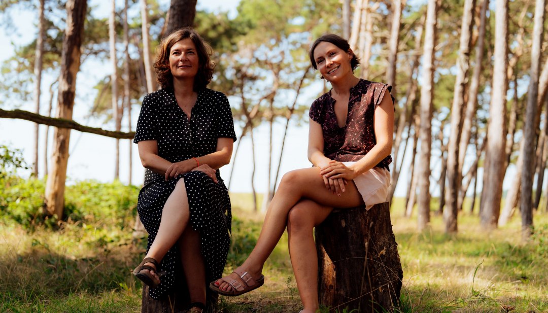 Dwie kobiety siedzą na pniach drzew w jasnym lesie nadmorskim, a w tle mienią się wydmy i morze. // Anne Liedtke i Maria Decker są zakochane w wyspie Rugii – i chcą się tym podzielić. // © MV-T/Petermann Dwie kobiety siedzą na pniach drzew w jasnym lesie nadmorskim, a w tle mienią się wydmy i morze.