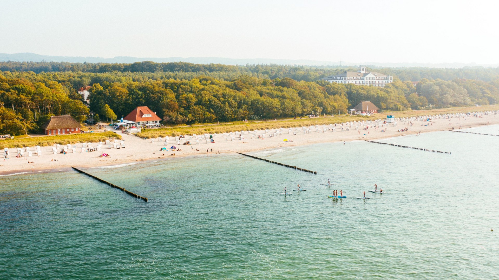 Luchtfoto van het strand in Kühlungsborn met strandstoelen, badgasten en stand-up paddlers tegen een groene bosrijke achtergrond.
