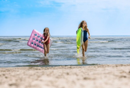 Dwoje dzieci wybiega z wody na plażę z dmuchanymi materacami pod pachami