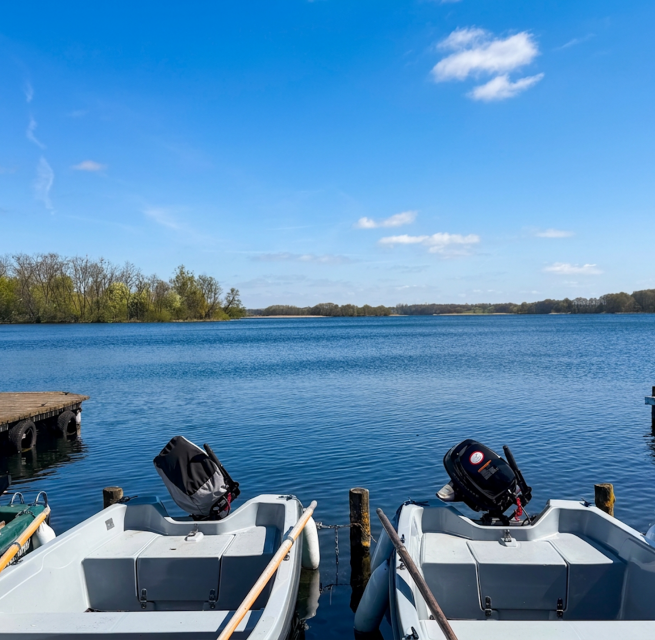 A view from the boat dock across Lake Krakow in bright sunshine. In the foreground, several rowboats and motorboats from the Kiefernhain vacation resort&rsquo;s boat rental service are moored at the dock. In the background, the vast, calm expanse of water stretches out, flanked by wooded shores. // &copy; Feriensiedlung Kiefernhain