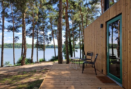 Houten terras van een klein huisje met uitzicht op het meer, omringd door pijnbomen in een rustige natuurlijke omgeving.