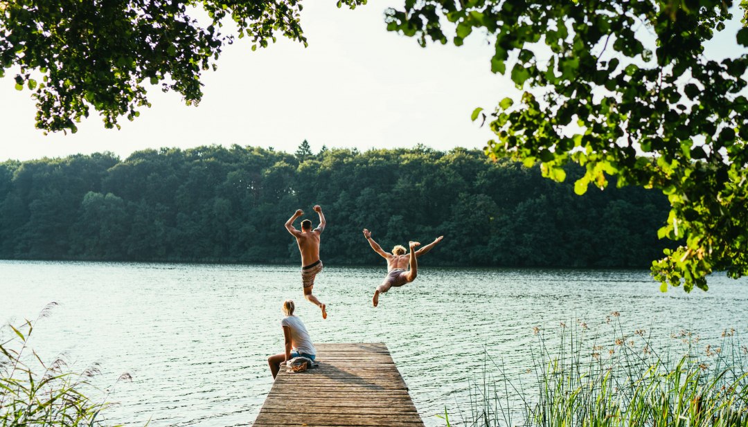 twee mensen die op een zomerse dag blij van een houten loopbrug in een zwemmeer springen omringd door bomen.