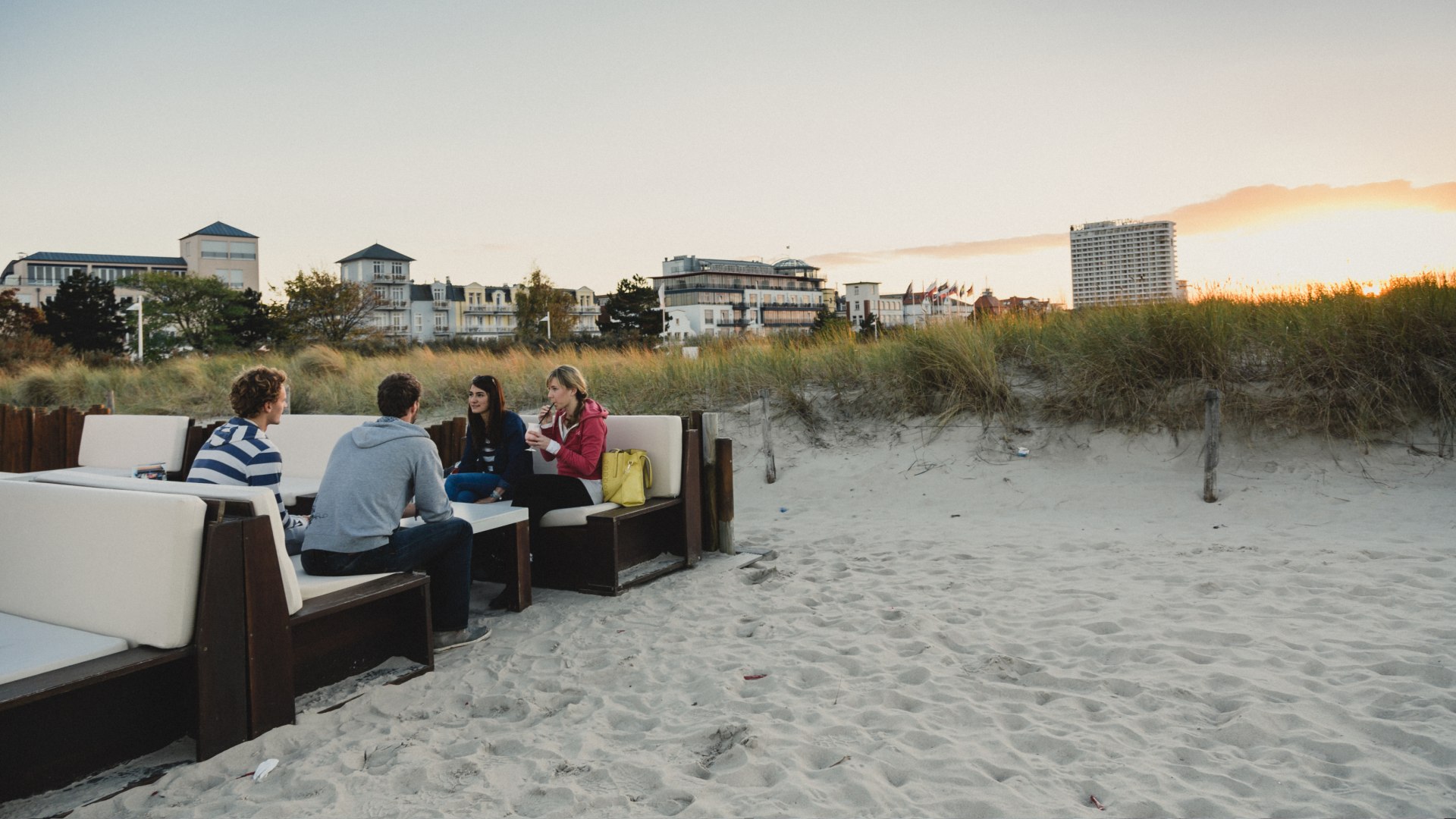 Sundowner with Baltic Sea flair - at Schuster's Warnemünde beach bar, relaxed beach enjoyment meets cozy lounge furniture and a maritime lifestyle right in the dunes., © TMV/Roth Group of young people sitting in lounge furniture on Warnemünde beach at sunset in front of Schuster's beach bar.