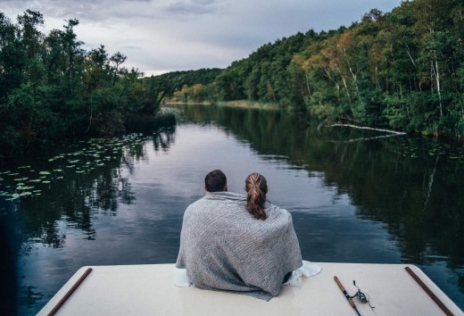 Een koppel zit op een woonboot, gewikkeld in een deken, uitkijkend over een rustige, beboste rivier in het Mecklenburgse merengebied.