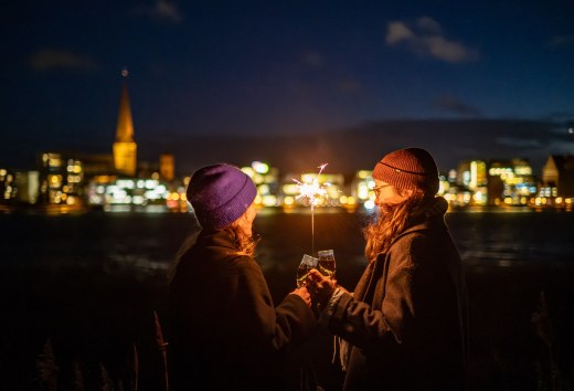 Twee mensen proosten met mousserende wijn terwijl ze een sparkler vasthouden, met de verlichte skyline van Rostock in Mecklenburg-Vorpommern op de achtergrond.