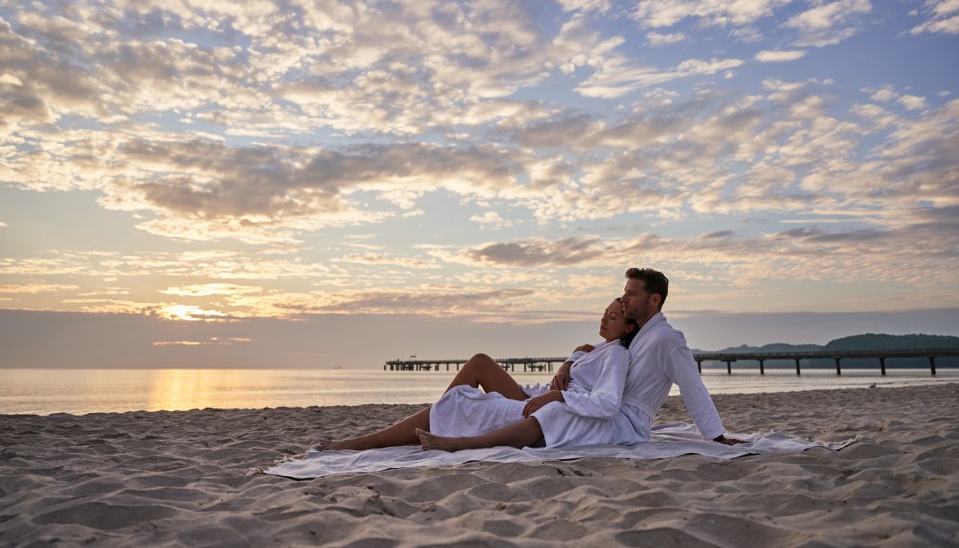 Two people sitting on the beach in bathrobes with the Binz pier in the background