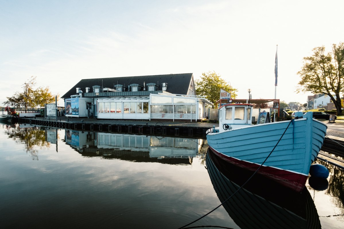 Restauracja Ribnitzer Fischhafen nad wodą: ł&oacute;dź przy pomostu, gładka jak lustro tafla wody &ndash; portowa atmosfera, kt&oacute;ra zachęca do odpoczynku. // &copy; Ribnitzer Fischhafen/Hugo Scholz-Witzel