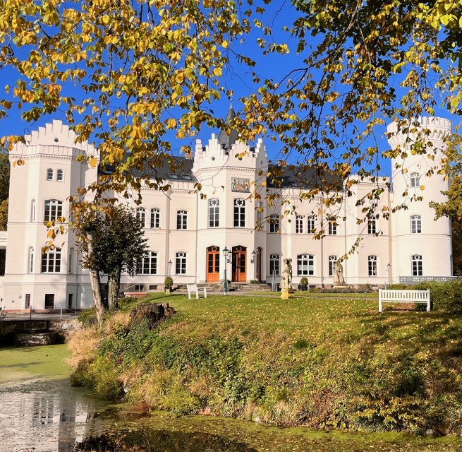 In front of Schlemmin Castle, autumn leaves glisten over the moat, and the white facade stands against a blue sky. // With its white castle facade, golden foliage, and tranquil moat, Schlemmin Castle showcases Mecklenburg-Western Pomerania at its finest. // © Schloss Schlemmin In front of Schlemmin Castle, autumn leaves glisten over the moat, and the white facade stands against a blue sky.