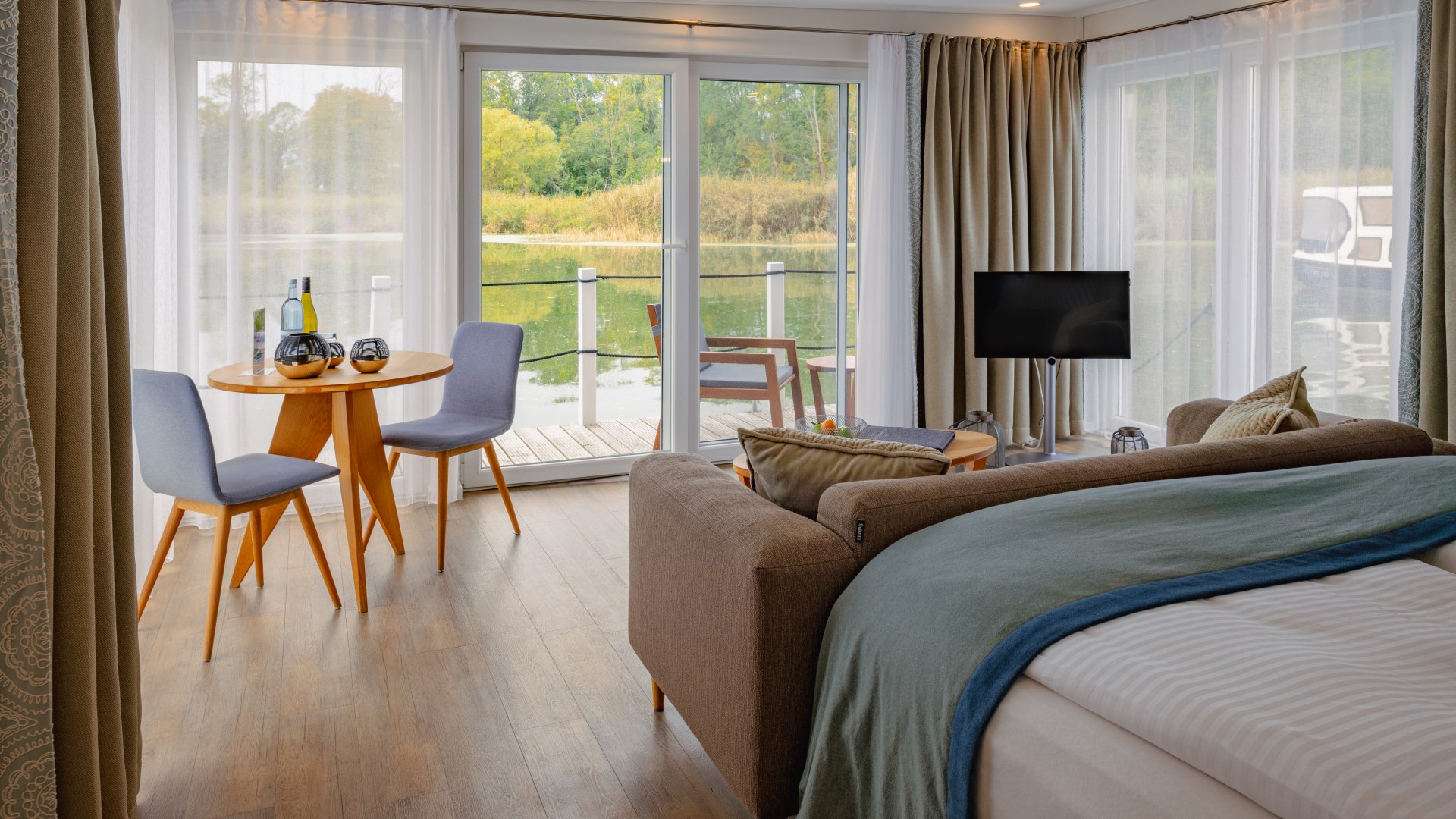 De drijvende suites in de natuurlijke haven van Krummin van binnen met bank, tv en uitzicht op het terras met zitje // Elegant interieur en ononderbroken uitzicht: De drijvende suites in de natuurlijke haven van Krummin // © MV-T/Tiemann De drijvende suites in de natuurlijke haven van Krummin van binnen met bank, tv en uitzicht op het terras met zitje