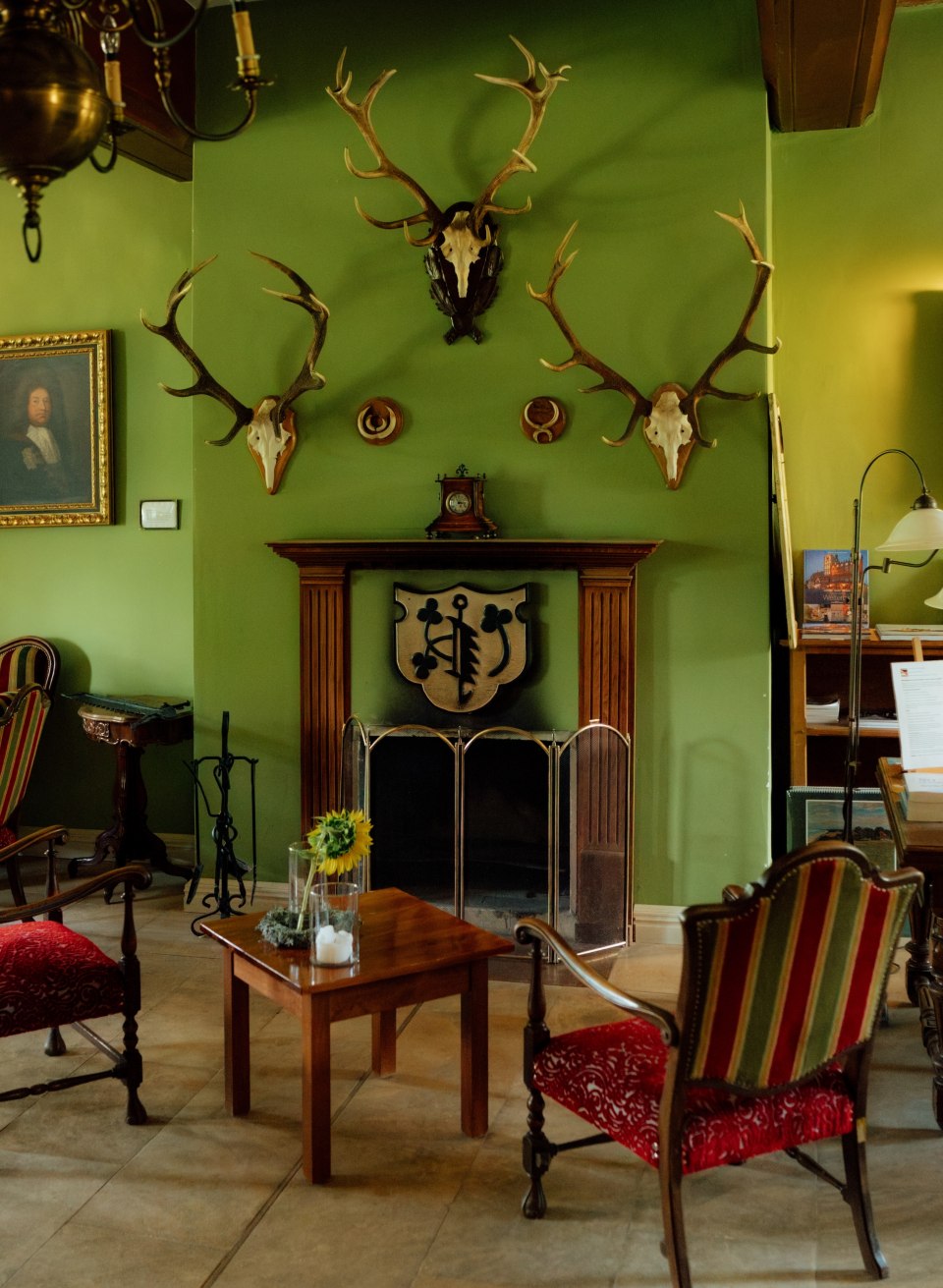 A fireplace with a wooden frame against a green wall, with antlers and a coat of arms above it; in front of it stand an armchair and a table in the Ludorf manor house. // Green-painted walls, old antlers, and a fireplace as the focal point: At the Ludorf Manor House, a space awaits where you can unwind—a place to feel at home in Mecklenburg-Western Pomerania. // © MV-T/Petermann A fireplace with a wooden frame against a green wall, with antlers and a coat of arms above it; in front of it stand an armchair and a table in the Ludorf manor house.