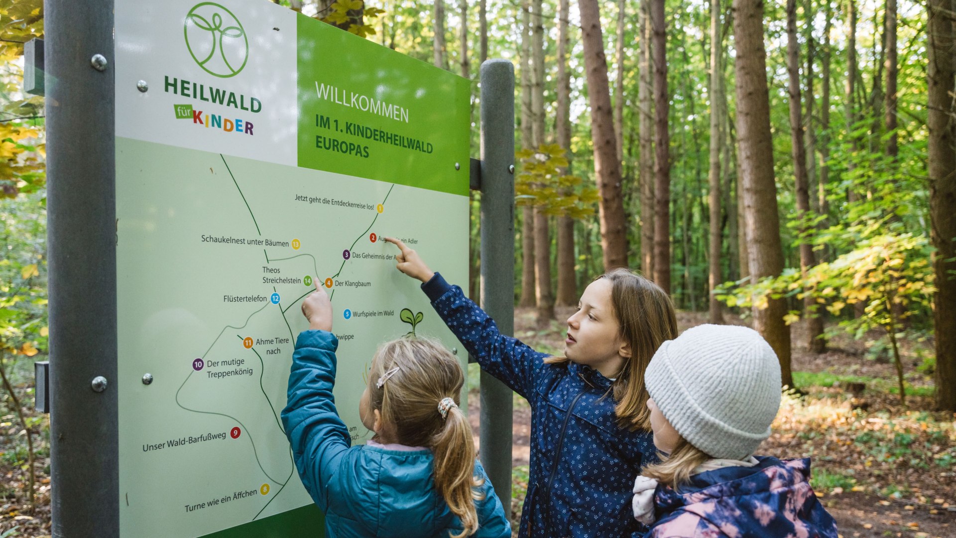 Drie kinderen staan voor een grote overzichtskaart in het bos, wijzen met hun vingers verschillende punten aan en plannen hun route.
