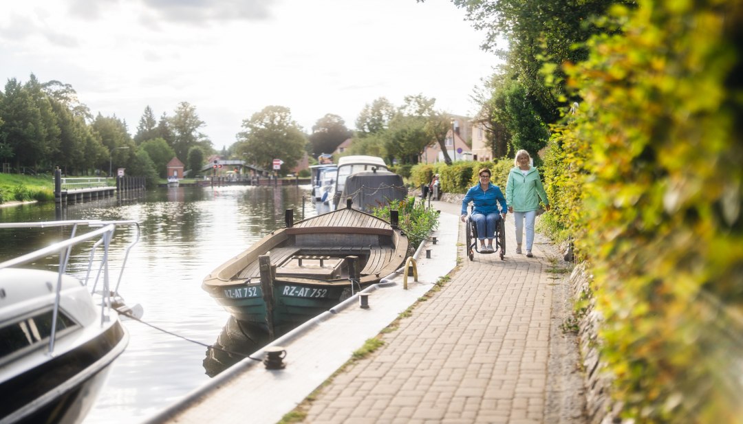 A wheelchair user out and about with her friend on the canal in Plau am See. Boats are moored at the edge.