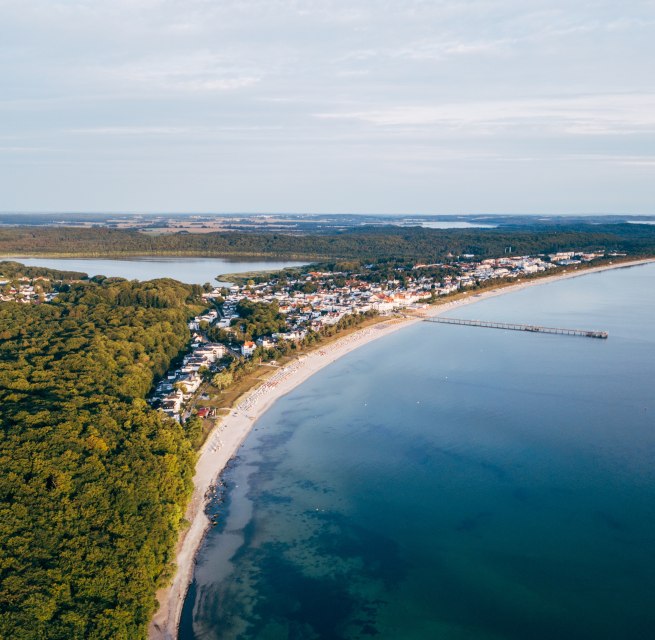 Widok z lotu ptaka na Binz na Rugii z molo, linią brzegową i otaczającymi lasami. // Widok z lotu ptaka na nadbałtycki kurort Binz na wyspie Rugia, który rozciąga się wzdłuż zakrzywionej linii brzegowej. Molo wystaje do spokojnej, turkusowej wody, a miasto otoczone jest gęstymi lasami i jeziorami. Niebo jest czyste, a spokojny krajobraz zachęca do odpoczynku. // © MV-T/Friedrich Widok z lotu ptaka na Binz na Rugii z molo, linią brzegową i otaczającymi lasami.