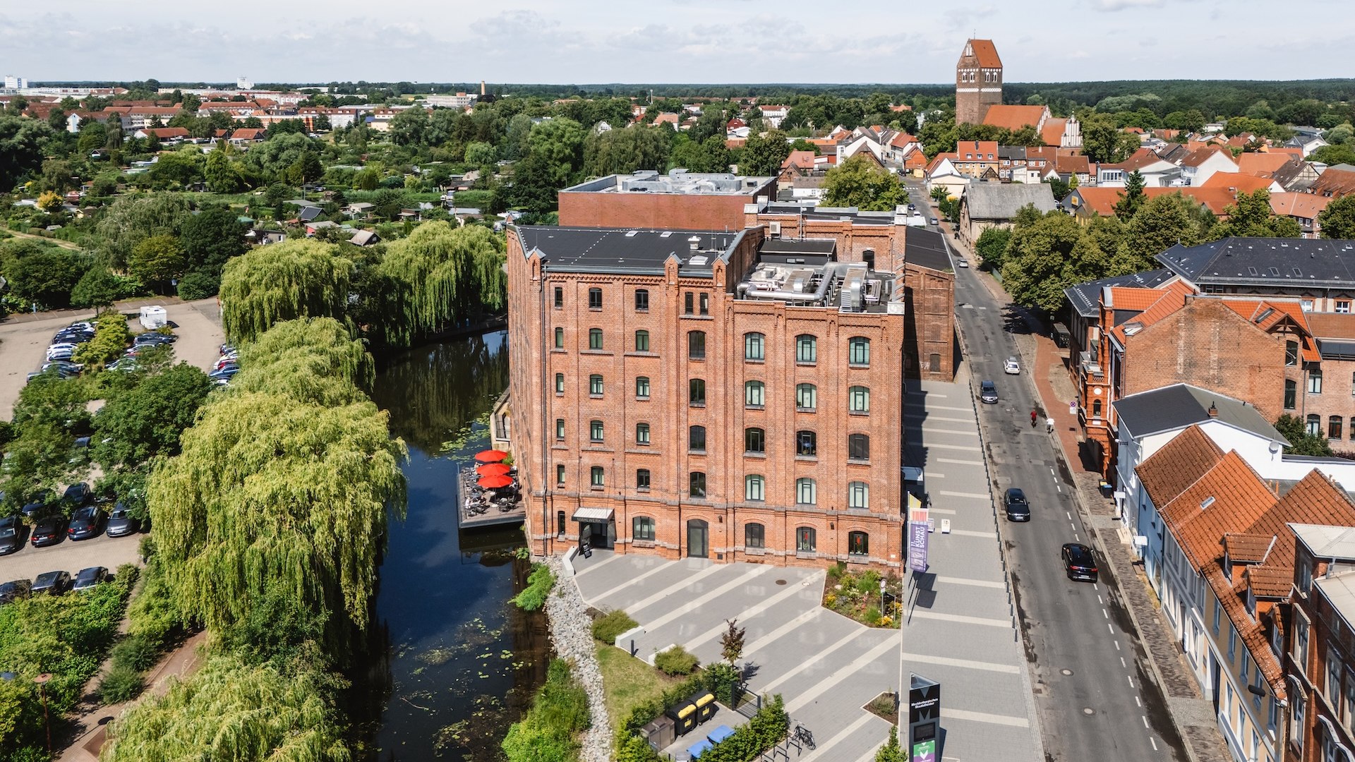 Een luchtfoto toont de Kulturm&uuml;hle in Parchim met zijn historische gevel en een gezellig restaurantterras aan het water, omgeven door groen.
