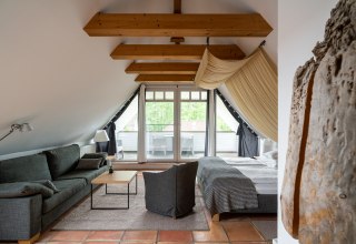 A stylishly furnished attic room with exposed wooden beams and a cozy ambience. The room offers a comfortable seating area, a large bed and access to a balcony with a view of the greenery. Warm terracotta floors and light fabrics round off the cozy design and invite you to relax., &copy; Hotel Carpe Diem
