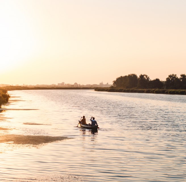 Two people paddle in a canoe across a calm river at sunset, with golden light on the water.