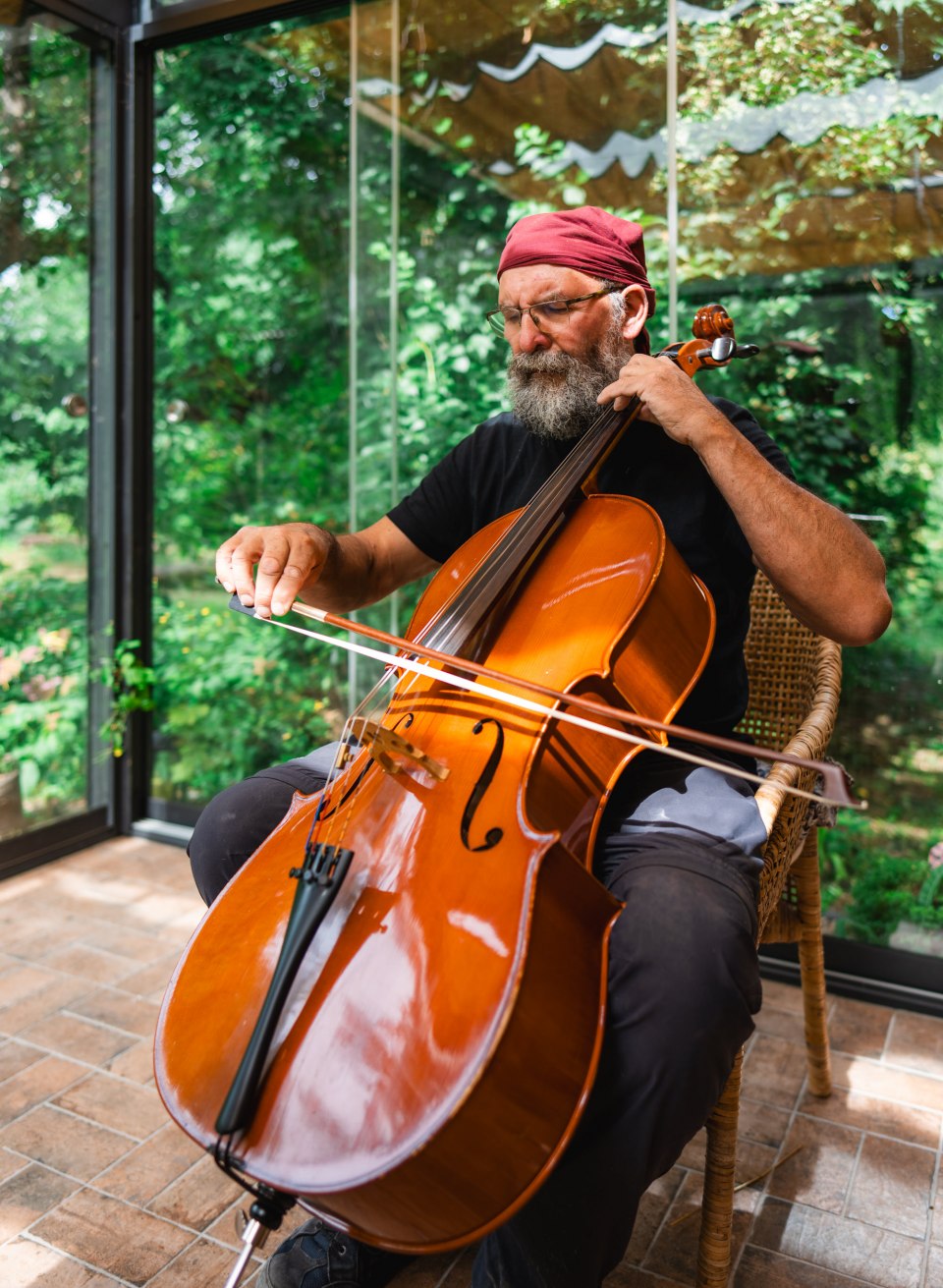 A man is playing the cello on a chair in the sunroom, focused and calm, surrounded by the green glow of the garden lights.