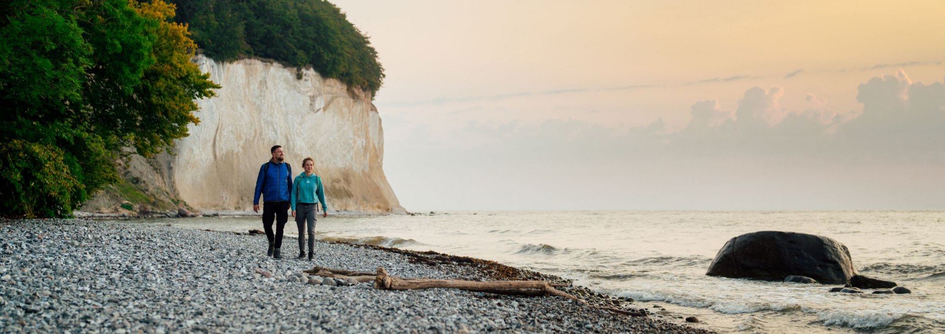 Couple walking along a pebble beach with chalk cliffs and gentle waves at sunset.
