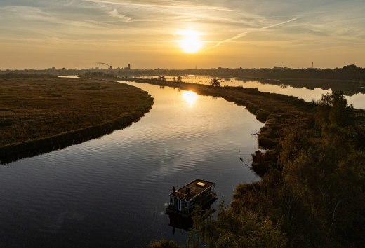 Woonboot op de Peene bij zonsondergang, omgeven door een vredig rivierlandschap en gouden licht aan de horizon.