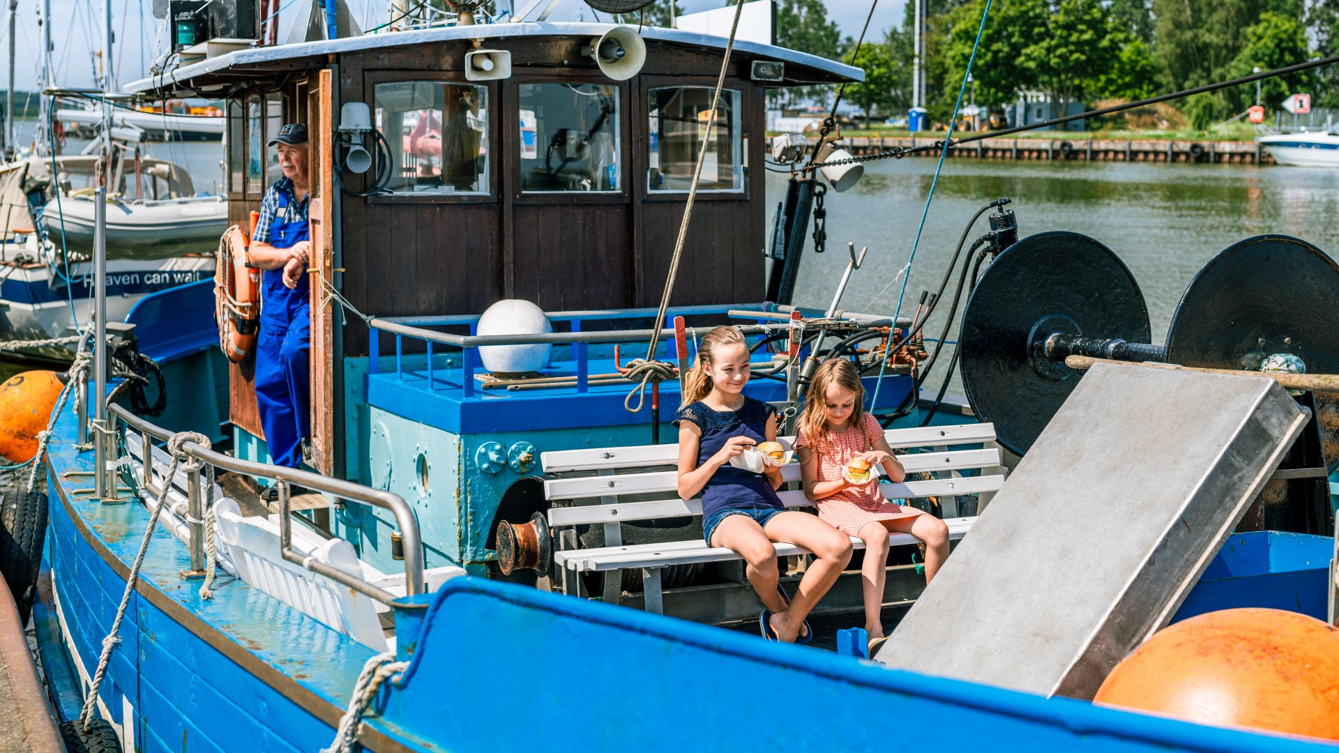 Twee kinderen eten een broodje vis op een vissersboot die normaal uit vissen gaat. De kapitein staat in de deur van de stuurhut.